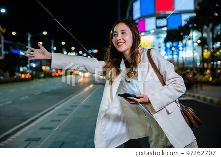 Beautiful woman smiling using smartphone application hailing with hand up calling cab after late work, Asian businesswoman standing hail waving hand taxi on road in busy city street at night Beautiful woman smiling using smartphone application hailing with hand up calling cab after late work, Asian businesswoman standing hail waving hand taxi on road in busy city street at night 97262707