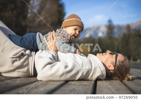 Happy family. Young mother playing with her baby boy infant oudoors on sunny autumn day. Portrait of mom and little son on wooden platform by lake. Positive human emotions, feelings, joy Happy family. Young mother playing with her baby boy infant oudoors on sunny autumn day. Portrait of mom and little son on wooden platform by lake. Positive human emotions, feelings, joy 97269050