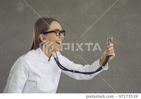 Profile view of a young smiling female doctor or nurse holding out a stethoscope and listening. 97273539