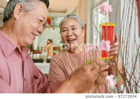 Joful senior couple decorating peach tree at home for Tet celebration Joful senior couple decorating peach tree at home for Tet celebration 97273934