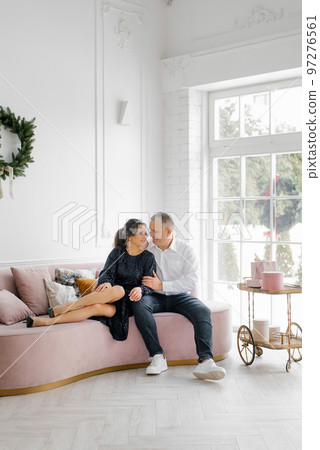 Happy lovers are sitting on a pink sofa in the living room, decorated for the New Year, they are hugging and celebrating the New Year Happy lovers are sitting on a pink sofa in the living room, decorated for the New Year, they are hugging and celebrating the New Year 97276561