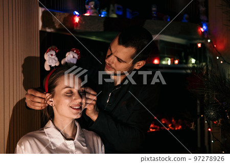 Merry Christmas and happy new year. Young couple in love celebrating Christmas or new year eve at home. Romantic couple in pajamas and Christmas reindeer antler headband hugging at home. 97278926