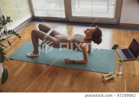 Slim woman doing exercises for legs with elastic band by online tutorials on laptop in living room 97289062