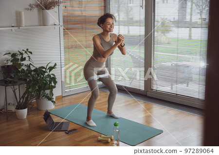 Slim woman doing exercises for legs with elastic band by online tutorials on laptop in living room Slim woman doing exercises for legs with elastic band by online tutorials on laptop in living room 97289064