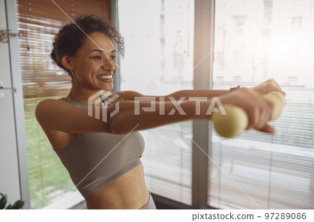 Smiling woman with dumbbells doing exercising at home and working on arms strength Smiling woman with dumbbells doing exercising at home and working on arms strength 97289086