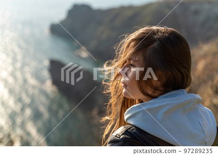 Woman tourist enjoying the sunset over the sea mountain landscape. Sits outdoors on a rock above the sea. She is wearing jeans and a blue hoodie. Healthy lifestyle, harmony and meditation 97289235