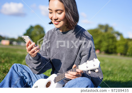 Technology and music. Smiling asian hipster girl sitting in park with smartphone and holding ukulele, playing instrument 97289435