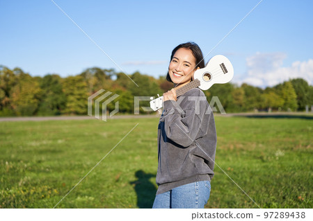 Young hipster girl, traveler holding her ukulele, playing outdoors in park and smiling Young hipster girl, traveler holding her ukulele, playing outdoors in park and smiling 97289438