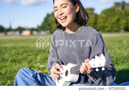 Portrait of beautiful asian woman singing, playing ukulele guitar in park, sitting alone on grass on sunny day 97289582