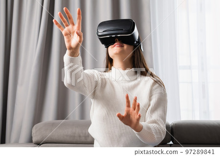 A beautiful caucasian woman uses VR headset touching an invisible screen sitting in the living room.  97289841