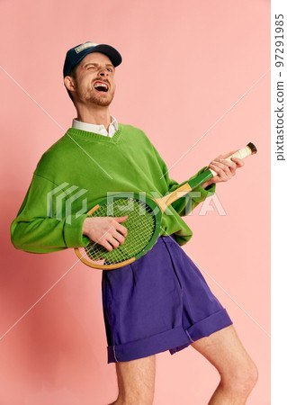 Portrait of young man in vintage sportive clothes playing tennis racket like guitar isolated over pink background. 97291985