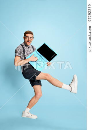 Young man in checkered shirt, shorts and suspenders playing with laptop like guitar isolated over blue background. Working break 97292289
