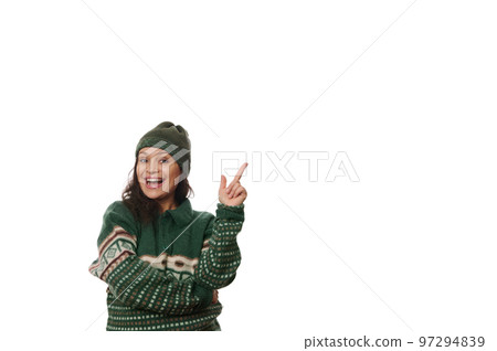 Beautiful dark-haired curly woman, in Xmas patterned warm green sweater and a wool hat, gesturing, pointing with her index finger at copy space, expressing positive emotions, smiling looking at camera 97294839