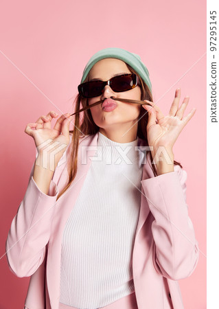 Portrait of young beautiful girl in a suit, hat and sunglasses posing over pink background. Good mood 97295145