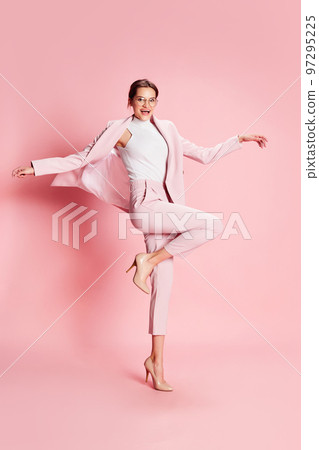 Portrait of beautiful young girl in suit and glasses posing on heels over pink background. Positive, good mood Portrait of beautiful young girl in suit and glasses posing on heels over pink background. Positive, good mood 97295225