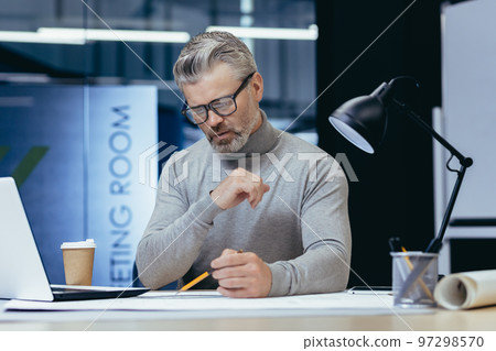 Concentrated senior male architect working in the evening by a lamp in the office. Sitting at the table, drawing a plan with a pencil, working with papers. 97298570