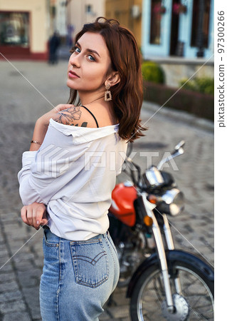 Young beautiful woman shows her bare shoulder with a tattoo standing by a bike on the street 97300266