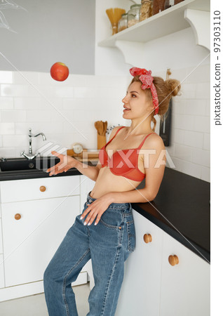 Young slim woman in lingerie bra and jeans with an apple at home kitchen Young slim woman in lingerie bra and jeans with an apple at home kitchen 97303110