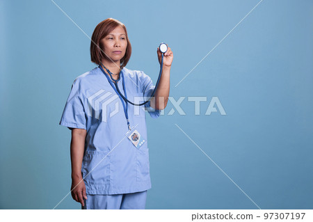 Elderly asian nurse in blue uniform holding medical stethoscope used for checkup consultation. Serious practitioner assistant working at healthcare facility, medicine service and concept Elderly asian nurse in blue uniform holding medical stethoscope used for checkup consultation. Serious practitioner assistant working at healthcare facility, medicine service and concept 97307197