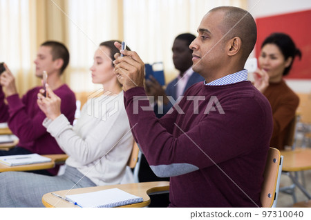 Students of different age sitting together at tables, shooting lesson on smartphones in classroom Students of different age sitting together at tables, shooting lesson on smartphones in classroom 97310040