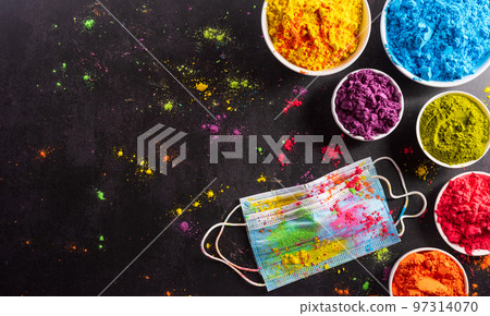 Top view of colorful holi powder with multicolored medical mask holi paint on dark background. Happy holi festival during coronavirus . (Covid-19) pandemic concept. 97314070