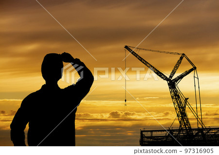 Silhouettes of cranes and workers towering over a construction site against the backdrop of the setting sun 97316905