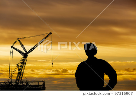 Silhouettes of cranes and workers towering over a construction site against the backdrop of the setting sun Silhouettes of cranes and workers towering over a construction site against the backdrop of the setting sun 97316907