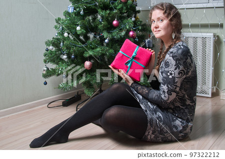 A woman sits on the floor next to a Christmas tree and holds a present in her hands 97322212