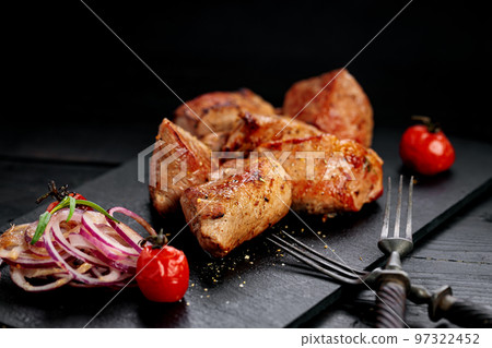 Appetizing pork shish kebab on a black stone cutting board. Red onion, cherry tomatoes and two forks. Soft focus 97322452