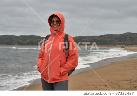 Smiling woman in an orange tourist jacket with a hood on the shore of the cold northern sea 97322487