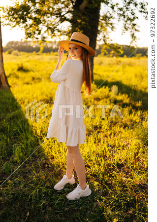 Vertical portrait of gorgeous redhead young woman in straw hat and white dress standing posing on beautiful meadow of green grass looking at camera, on background of warm sunlight at summer day. Vertical portrait of gorgeous redhead young woman in straw hat and white dress standing posing on beautiful meadow of green grass looking at camera, on background of warm sunlight at summer day. 97322692