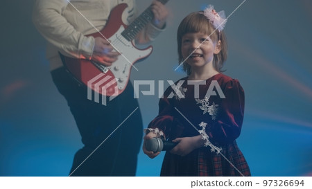 Little girl on stage in vintage dress, she sings into microphone and dances, her father plays an electric guitar. Color music is shining and smoke is billowing. Family time, creativity and hobbies Little girl on stage in vintage dress, she sings into microphone and dances, her father plays an electric guitar. Color music is shining and smoke is billowing. Family time, creativity and hobbies 97326694