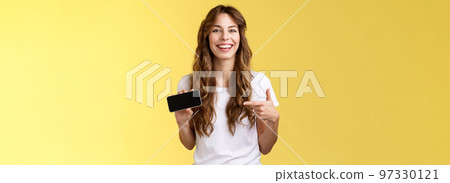 Carefree charismatic cute curly-haired female gamer likes playing smartphone games showing own game score hold mobile phone horizontal pointing display laughing pleased amused 97330121