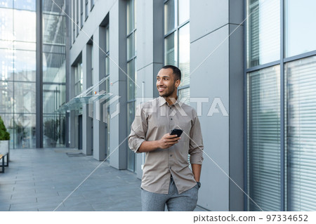 Smiling hispanic man walking down street near modern office building, freelancer businessman looking away holding mobile phone. Smiling hispanic man walking down street near modern office building, freelancer businessman looking away holding mobile phone. 97334652