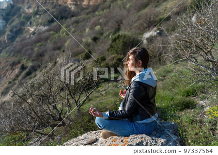 Woman tourist enjoying the sunset over the sea mountain landscape. Sits outdoors on a rock above the sea. She is wearing jeans and a blue hoodie. Healthy lifestyle, harmony and meditation Woman tourist enjoying the sunset over the sea mountain landscape. Sits outdoors on a rock above the sea. She is wearing jeans and a blue hoodie. Healthy lifestyle, harmony and meditation 97337564