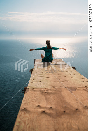 A girl sits on a wooden springboard for jumping with a rope. In a dark green dress with her hands in the air. 97337580