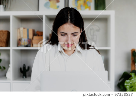 Brunette woman working with laptop at home or office. Girl is reading computer screen and smiling. Restrained joyful face. Blurred background with furniture. Person working from home concept 97338557