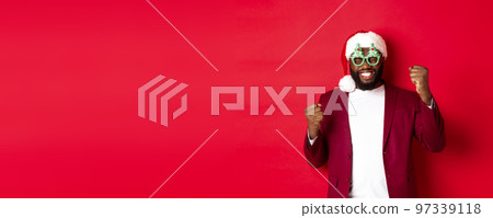 Merry Christmas. Cheerful Black man wearing funny party glasses and santa hat, smiling joyful, celebrating winter holidays, standing over red background Merry Christmas. Cheerful Black man wearing funny party glasses and santa hat, smiling joyful, celebrating winter holidays, standing over red background 97339118