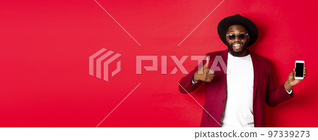 Handsome and stylish Black man showing phone screen and thumb up at camera, recommending online store app, red background 97339273
