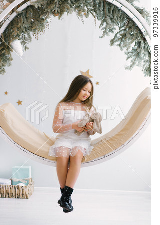 Cute girl holding a soft favorite toy and sitting on a swing decorated for Christmas Cute girl holding a soft favorite toy and sitting on a swing decorated for Christmas 97339916