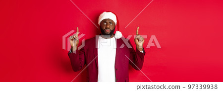Christmas, party and holidays concept. Upset african-american guy in santa hat complaining, pointing fingers up and frowning disappointed, standing against red background 97339938