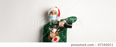 Concept of covid-19 and Christmas holidays. Happy guy in face mask and santa hat, celebrating New Year party and pouring glass of champagne, white background 97340051