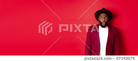 Fashion and party concept. Shocked and displeased black man staring at something unpleasant, looking upper left corner and frowning, standing against red background 97340079