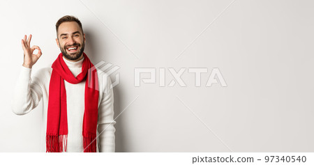 Winter holidays and shopping concept. Satisfied bearded man laughing and showing okay sign, approve and like product, standing in christmas scarf and sweater, white background Winter holidays and shopping concept. Satisfied bearded man laughing and showing okay sign, approve and like product, standing in christmas scarf and sweater, white background 97340540
