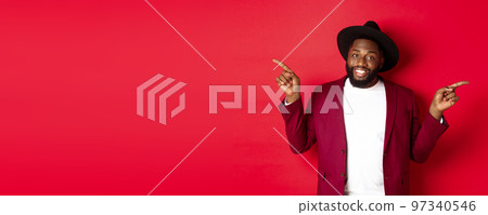 Winter holidays and shopping concept. Cheerful Black man smiling and showing two promo, pointing fingers sideways at copy spaces, red background Winter holidays and shopping concept. Cheerful Black man smiling and showing two promo, pointing fingers sideways at copy spaces, red background 97340546