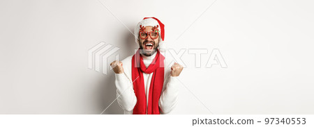Christmas, New Year and celebration concept. Excited man in Santa hat and party glasses, winning and rejoicing, making fist pump, shouting yes, standing over white background Christmas, New Year and celebration concept. Excited man in Santa hat and party glasses, winning and rejoicing, making fist pump, shouting yes, standing over white background 97340553