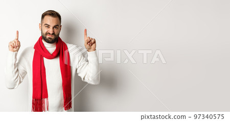 Christmas shopping and winter holidays concept. Displeased and skeptical guy complaining, pointing fingers up at bad product, standing against white background Christmas shopping and winter holidays concept. Displeased and skeptical guy complaining, pointing fingers up at bad product, standing against white background 97340575