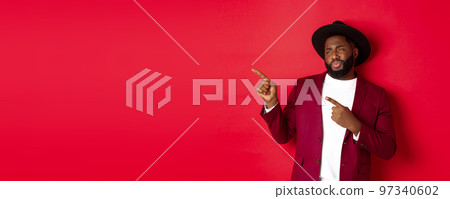 Winter holidays and shopping concept. Skeptical and displeased Black man looking with disdain and pointing finger left at copy space, standing against red background 97340602