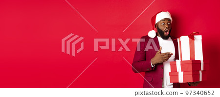 Christmas, New Year and shopping concept. Surprised Black man looking at xmas presents with amazement, wearing santa hat, standing with gifts against red background 97340652
