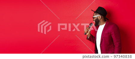 Passionate black male singer performing against red background, singing into microphone, wearing party outfit, standing over red background Passionate black male singer performing against red background, singing into microphone, wearing party outfit, standing over red background 97340838